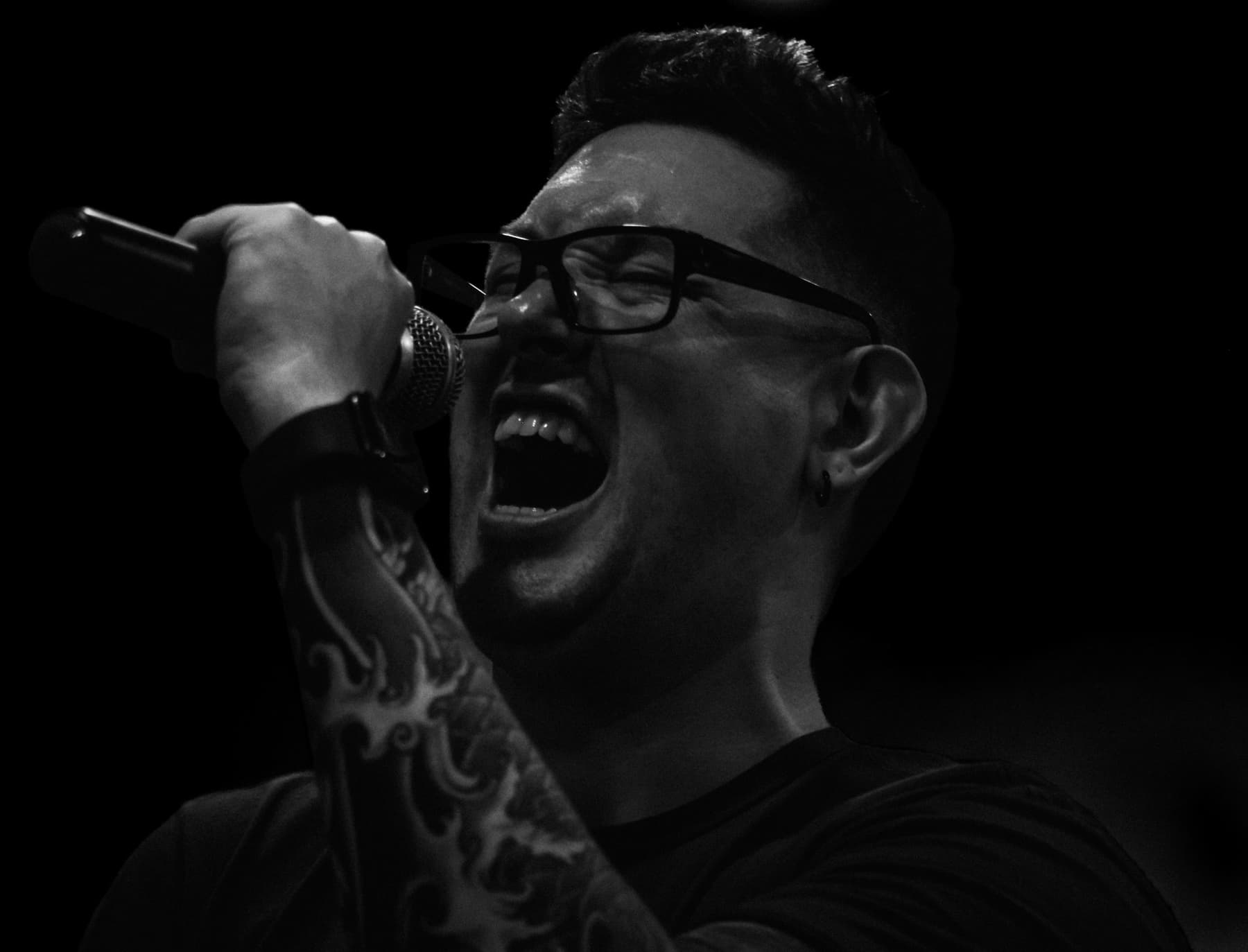 The voice of Conflict/Resolution mid-scream into a microphone, black and white, tattooed arm raised